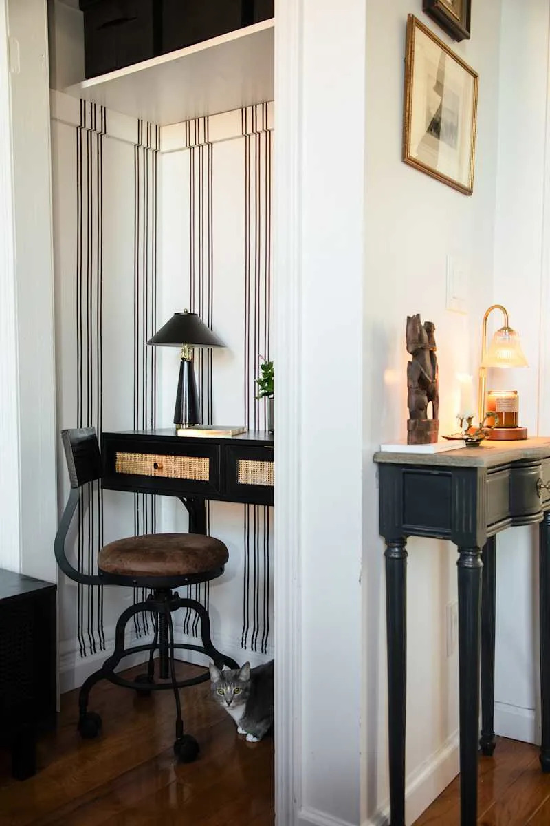 Cozy workspace with a black desk, a lamp, and a cat peeking from the corner, featuring striped wallpaper and wooden flooring.