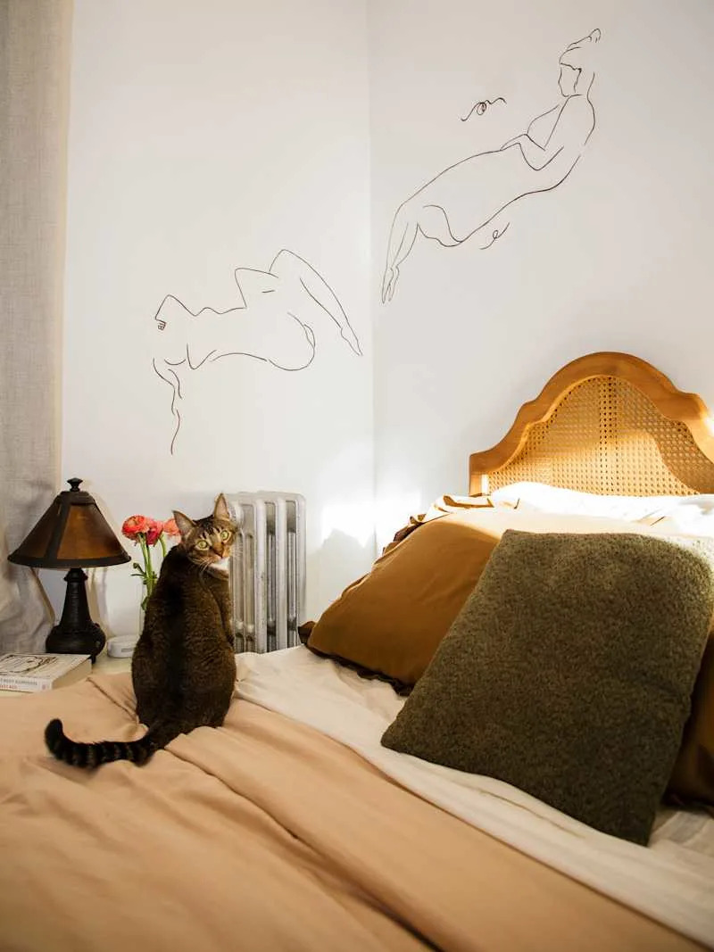 A cozy bedroom featuring a cat on a bed with brown and cream bedding, decorative pillows, and wall art of abstract figures.