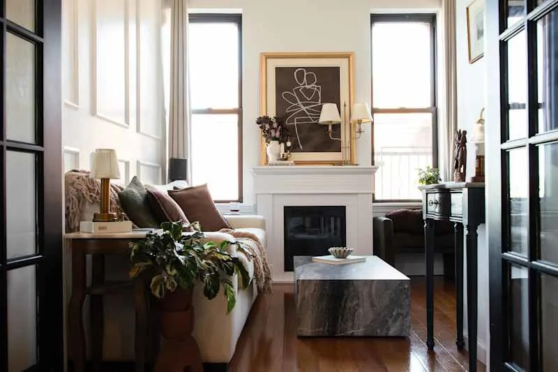 Cozy living room with a white sofa, brown pillows, a marble coffee table, and a fireplace, featuring large windows and artwork.