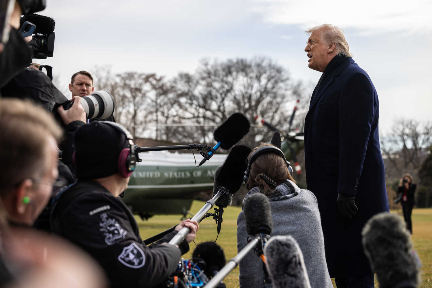 President Trump, wearing a long dark coat, speaks to the media. Behind him, a green helicopter is parked.
