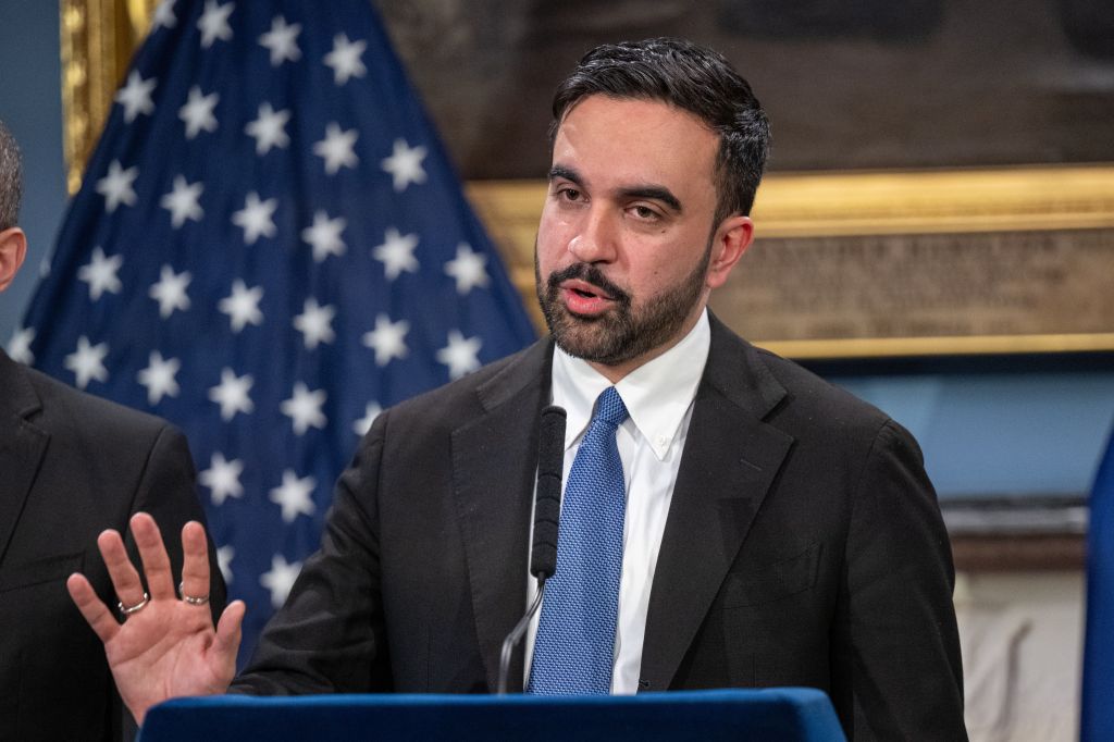 Zohran Mamdani speaking at a press conference with an American flag behind him.
