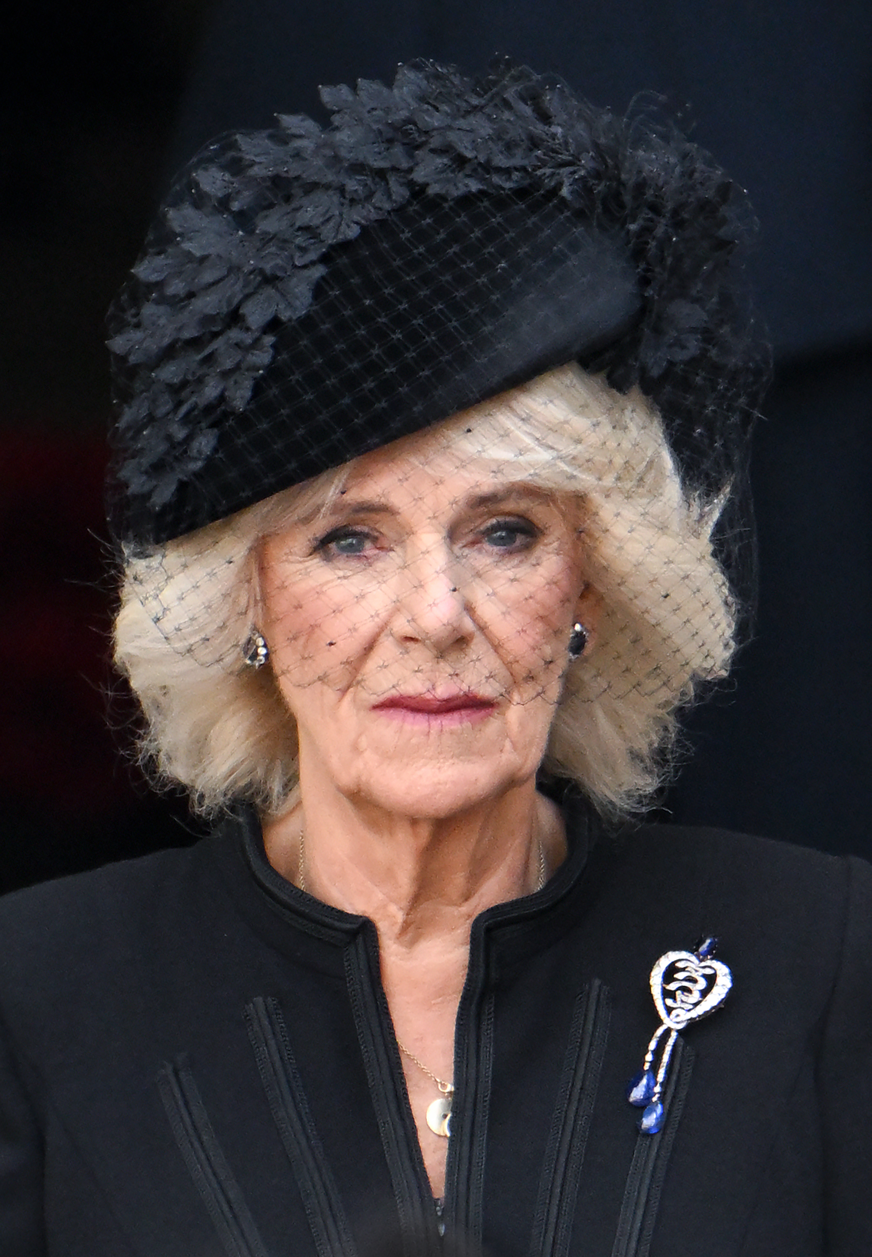 Queen Camilla wearing a black hat and coat with a heart brooch at Queen Elizabeth's funeral