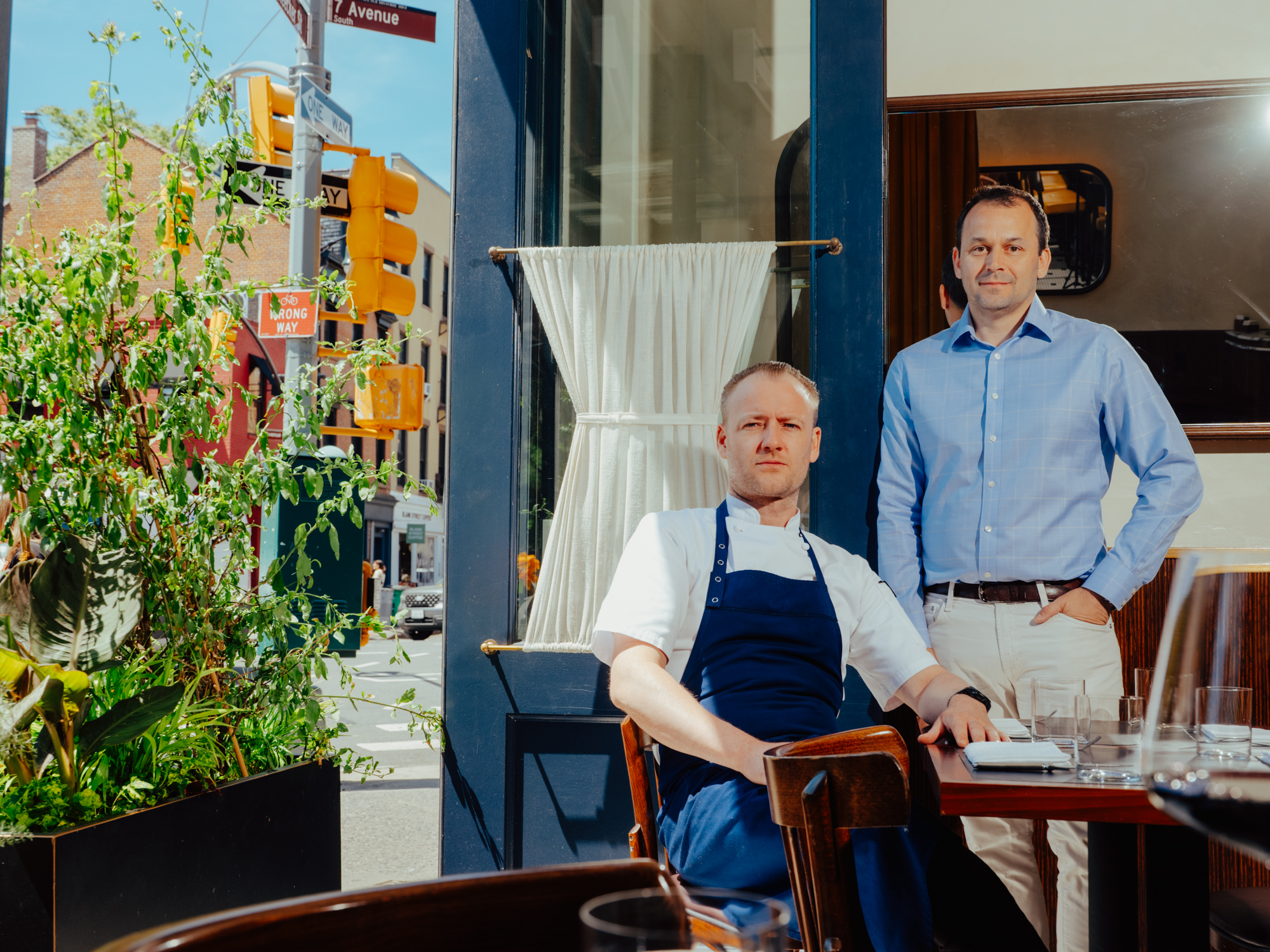 One man in an apron sitting at the table with another man in a blue shirt standing next to him.