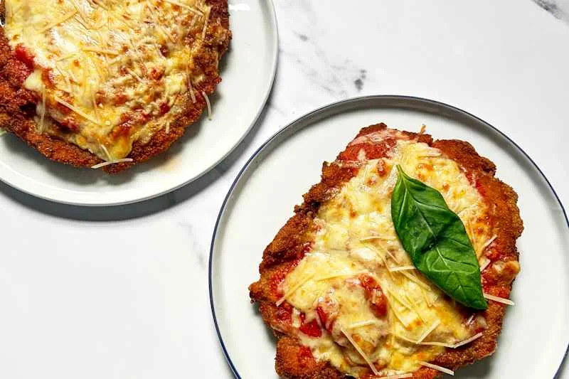 Two plates of chicken parmesan topped with melted cheese, marinara sauce, and fresh basil leaves.