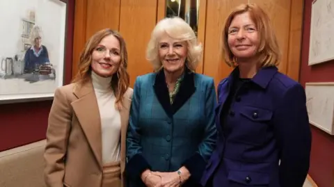 PA Media Geri Halliwell-Horner, the Queen and Maggie's chief executive Dame Laura Lee stand in a wood panelled booth. Horner-Halliwell wears a beige suit with a white turtle neck, the Queen wears a blue tweed suit with black colour and cuffs, Dame Laura is wearing a purple jacket and dark trousers. 