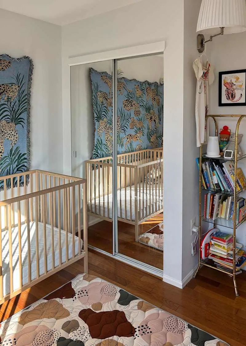 Nursery with a wooden crib, mirrored closet, colorful floral rug, and a bookshelf filled with children's books.