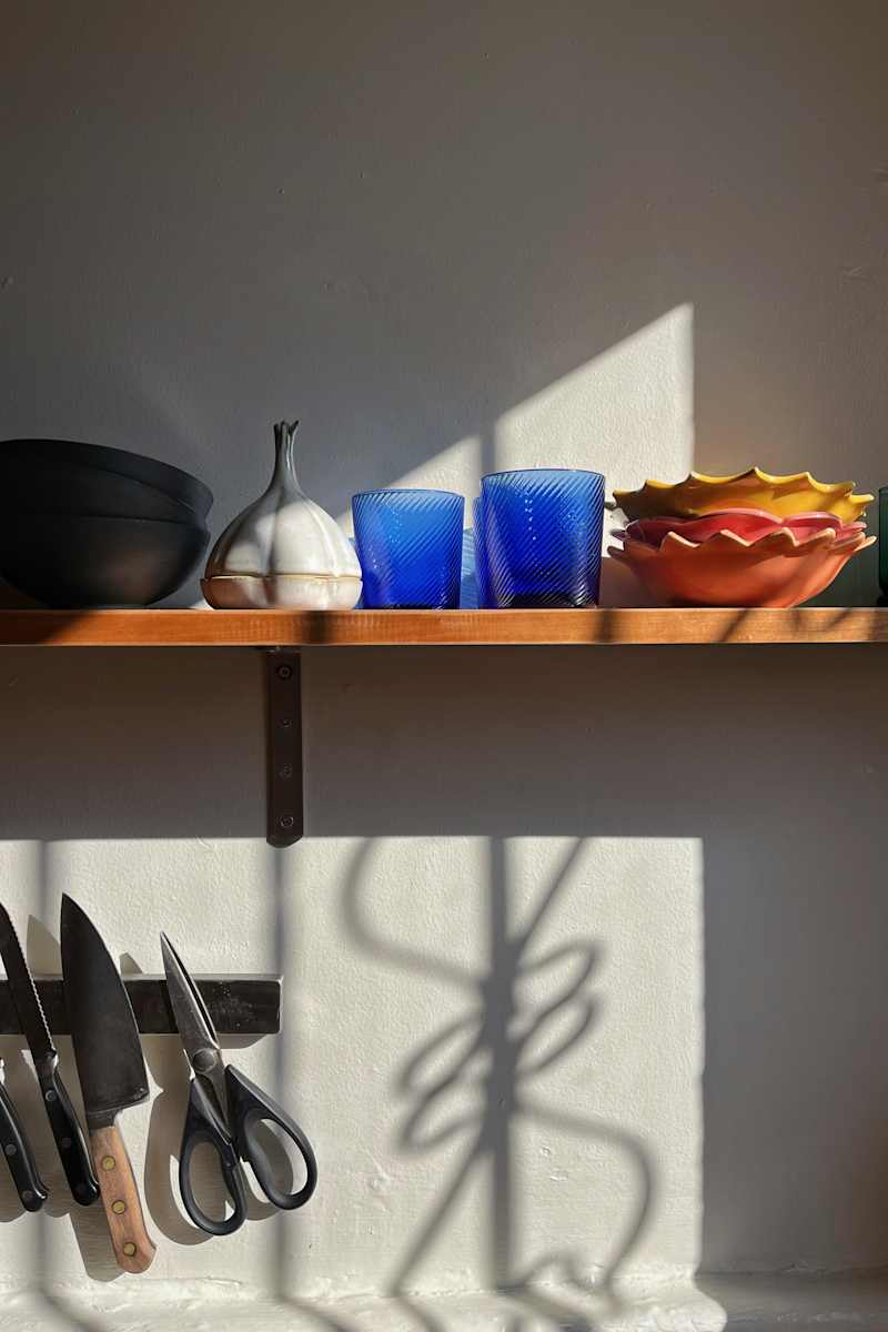 A wooden shelf displaying a black bowl, a white decorative piece, two blue glasses, and colorful bowls, with kitchen knives and scissors below.
