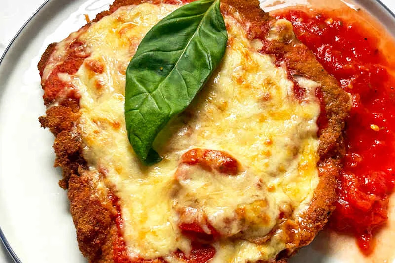 Breaded chicken topped with melted cheese and basil, served with marinara sauce on a white plate.
