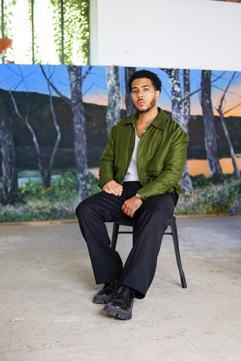 Individual seated on a chair in front of a painted landscape.