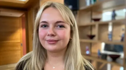 Caitlyn Warrington looks at the camera. She is in her early 20s and has shoulder length blonde hair. She is standing in a wood panelled toom with shelves holding ornaments in the background.