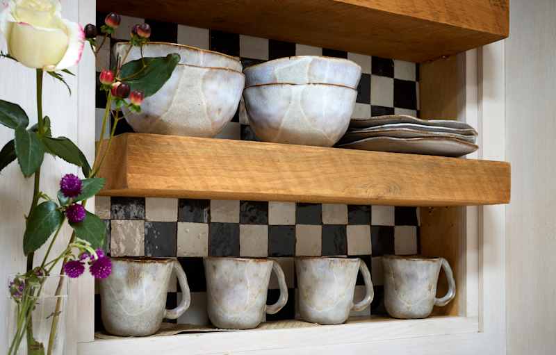 Ceramic bowls and mugs on wooden shelves, with a checkered tile background and a white rose nearby.