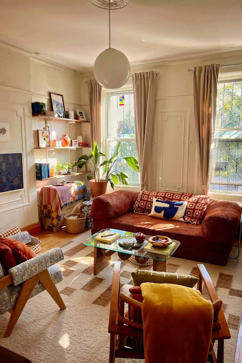 Cozy living room with a brown corduroy sofa, patterned pillows, a glass coffee table, and plants by the window.