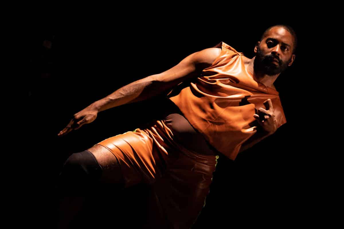 Alameda Post - Jerron, a dark-skinned Black man, leans with arm outstretched in a dark orange tunic.