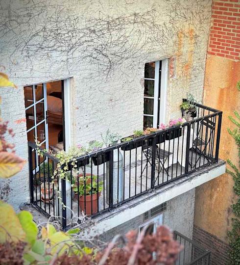 Balcony with black railing, potted plants, and a small table with chairs, overlooking a textured wall.