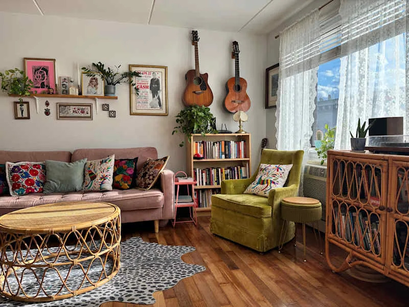 Cozy living room with a pink sofa, colorful pillows, a round rattan coffee table, green armchair, and bookshelves.