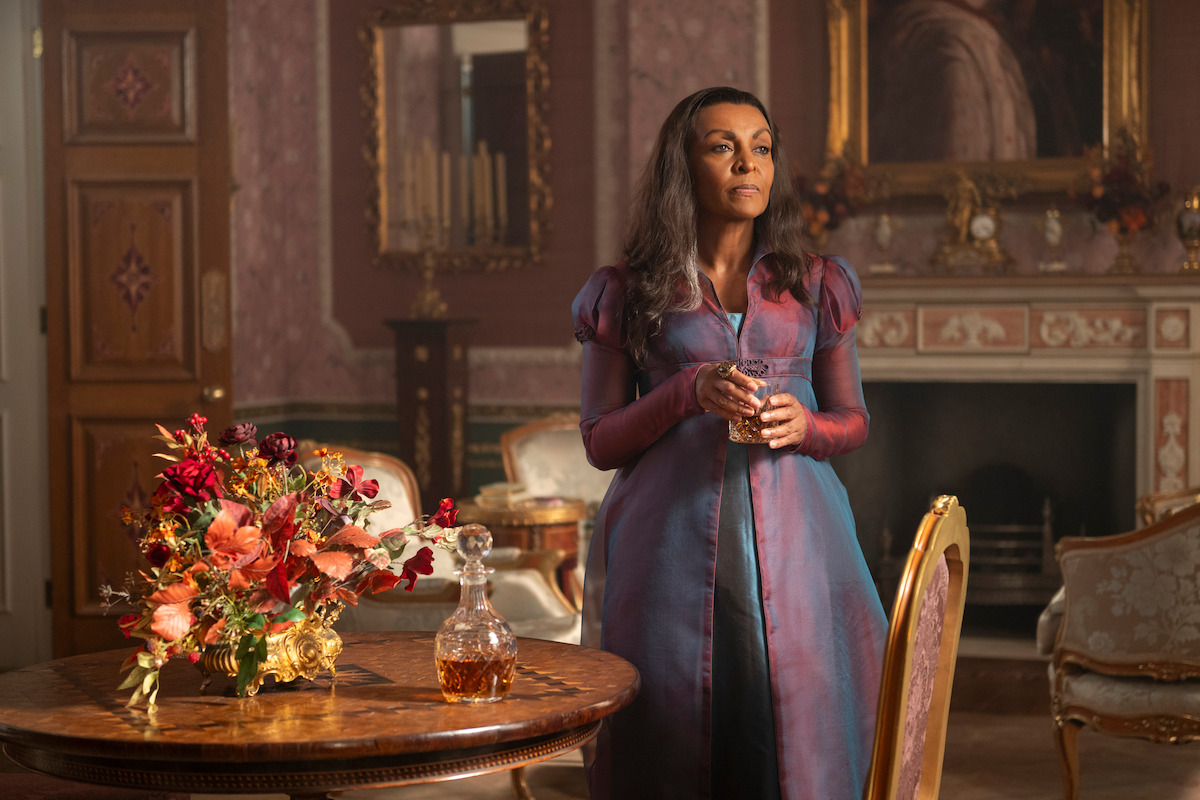 A woman in a purple period dress stands in an ornate, vintage parlor holding a glass. A floral bouquet and decanter sit on a round wooden table. The room has rich décor and a fireplace in the background.