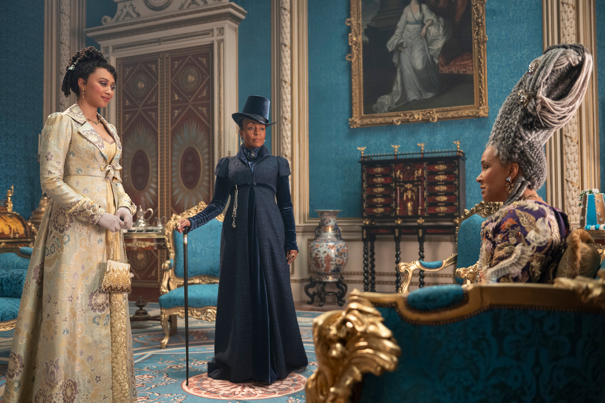 Three women in elaborate historical costumes converse in a lavish, ornate blue and gold room with royal decor and classical paintings, suggesting a regal or aristocratic setting.