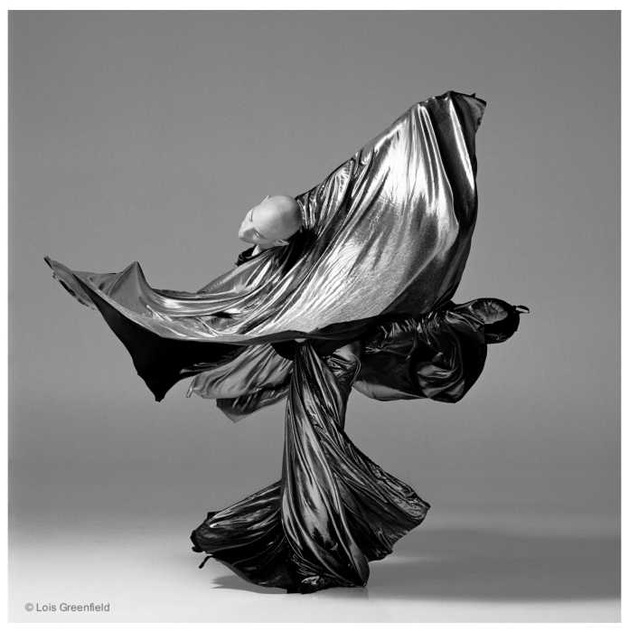 Dance Piece Untitled #1 After Martha Graham, circa 1994, photo by Lois Greenfield. A black-and-white photograph of Arthur Avilés captured mid-air, wearing a long, flowing dress that billows outward. The dancer’s legs are tucked beneath the fabric, arms curved—one overhead and one extended to the side. The head is tilted back, and the background transitions from light at the bottom to dark at the top.