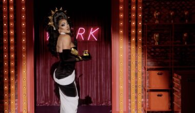 Photo of a drag queen wearing a black and white gown with a gold crown in front of a neon sign. Only the letters "R" and "K" are visible on the sign
