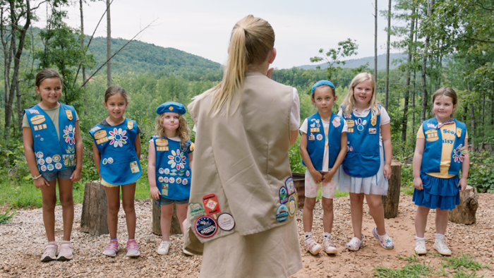 A group of young girls in blue uniforms with patches stand outdoors in a wooded area.