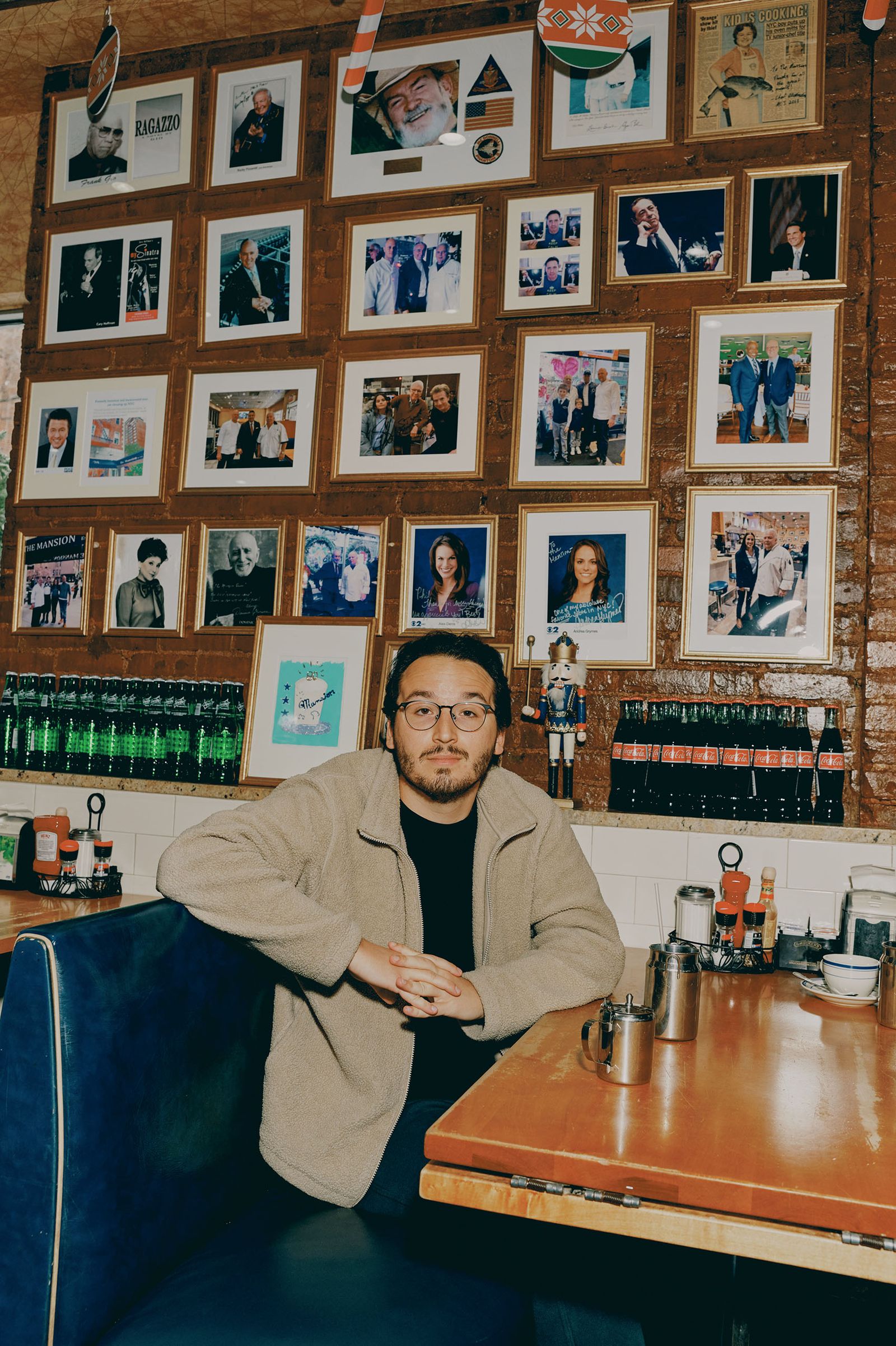 Figure sits at a booth in a restaurant setting. Behind him a wall is filled with photos of people.
