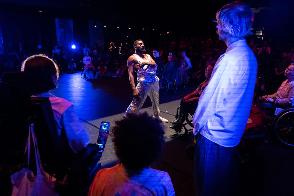 Alameda Post - Jerron, dressed in a silver jumpsuit underneath blue and purplish hues, stands in an aisle of light in front of an audience. 