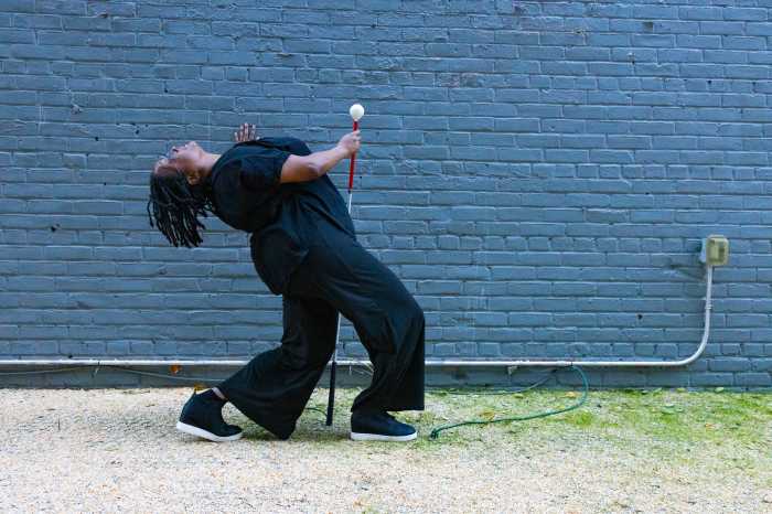 Against a gray brick wall, Kayla, a dark-skinned Black woman dressed in loose black clothing and black sneakers leans dramatically backward in a dance pose. Her knees are bent and her head tilts toward the sky, creating an arched shape with her body. In her right hand, she holds a white cane angled upward, which becomes part of the composition. Her left hand rests on her chest as if in gesture or emphasis. The ground beneath her is gravel, and a green extension cord trails across it.