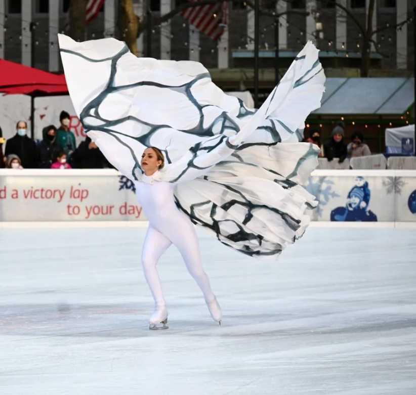 Figure Skater gliding along the ice in a white full body leotard and white cape with black design.