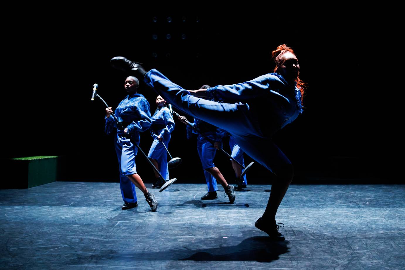 Dancers spotlit in blue jumpsuits. The foreground dancer in an open arabesque looking to the audience.