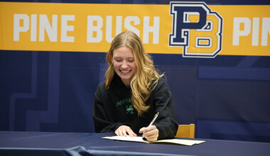 A high school girl with long blonde hair sits at a table, smiles and signs a piece of paper. Behind her is a banner that says Pine Bush PB.