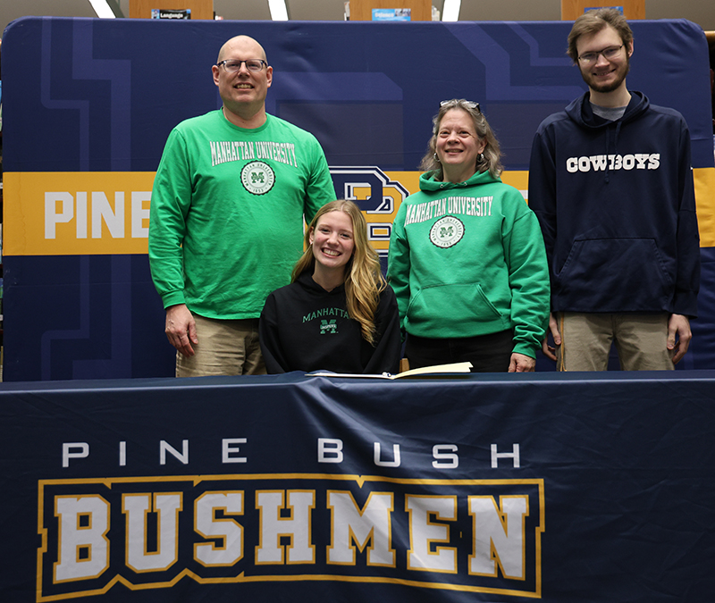 A high school senior with long blonde hair sits smiling at a table. Behind her are her family, two men and a woman.  Everyone is smiling