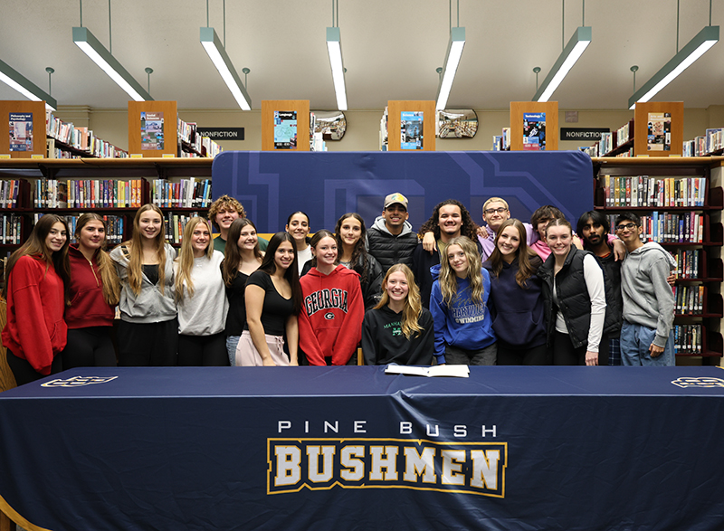 A large group of high school kids surround a girl who is sitting at a table smiling. They are all smiling.