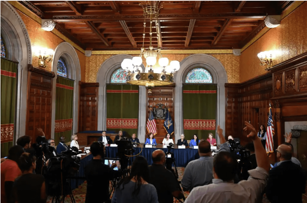 Interior government room with figures speaking in front of press 