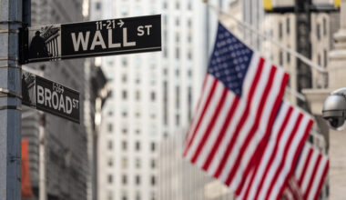 Wall street sign in New York City with American flags and New York Stock Exchange in background.