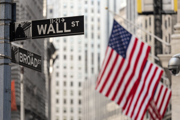 Wall street sign in New York City with American flags and New York Stock Exchange in background.