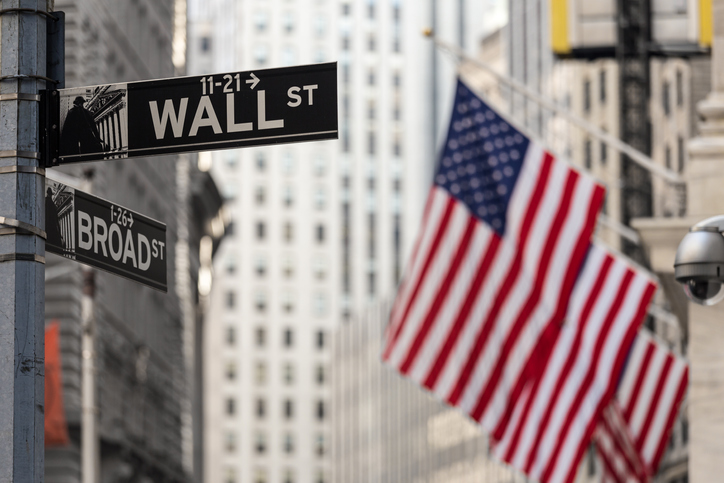 Wall street sign in New York City with American flags and New York Stock Exchange in background.