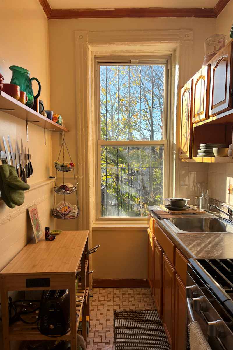 Cozy kitchen with wooden cabinets, a window overlooking trees, a bamboo cart, and kitchen utensils on display.