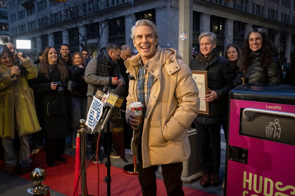 Andy Cohen smiles in a tan puffer coat holding a coffee cup, standing next to a hot pink "Landfill" trash can with "Hudson Square" written on its side.
