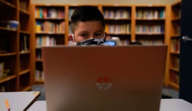 child, face covered by a mask, sits in front of a laptop in a library