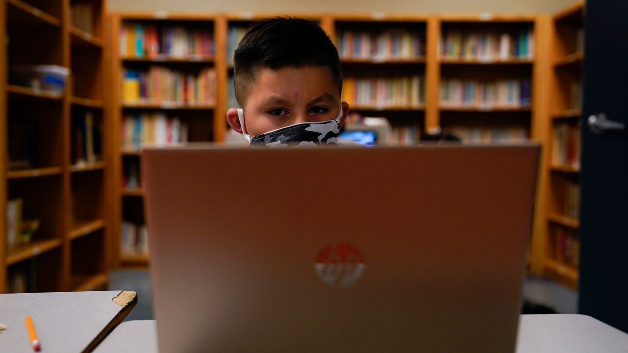 child, face covered by a mask, sits in front of a laptop in a library