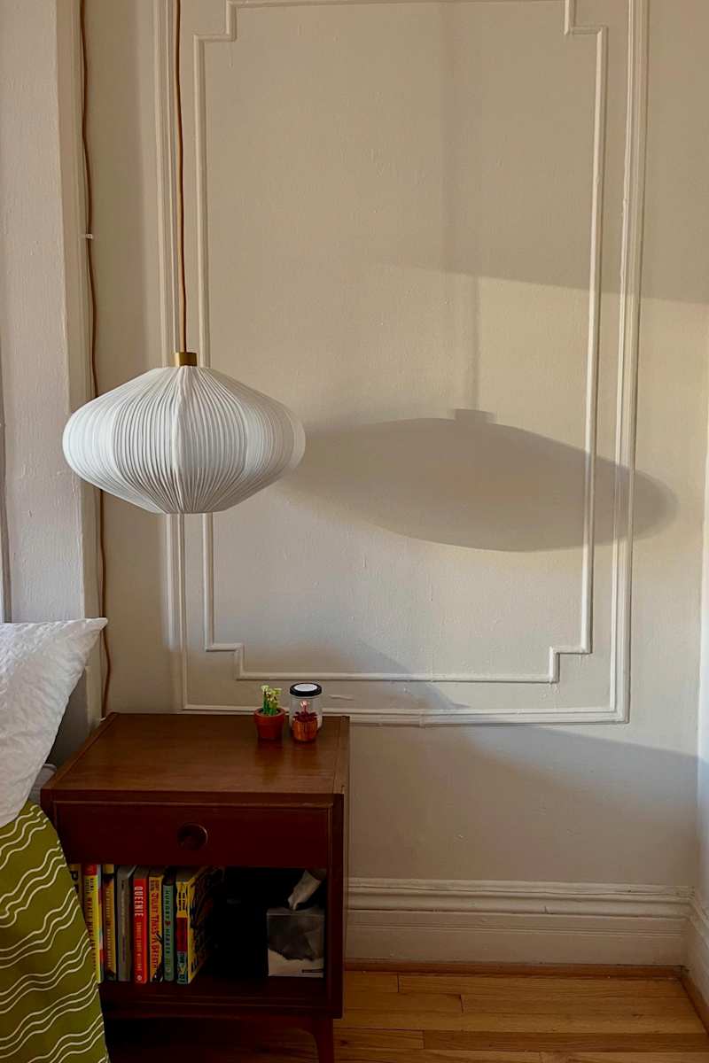 A white pendant lamp casts a shadow on a wall above a wooden nightstand with books, small plants, and a jar.