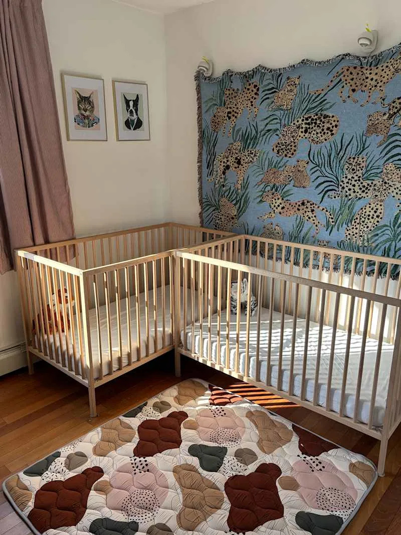 Nursery with two wooden cribs, a colorful animal-themed wall hanging, and a patterned play mat on wooden flooring.