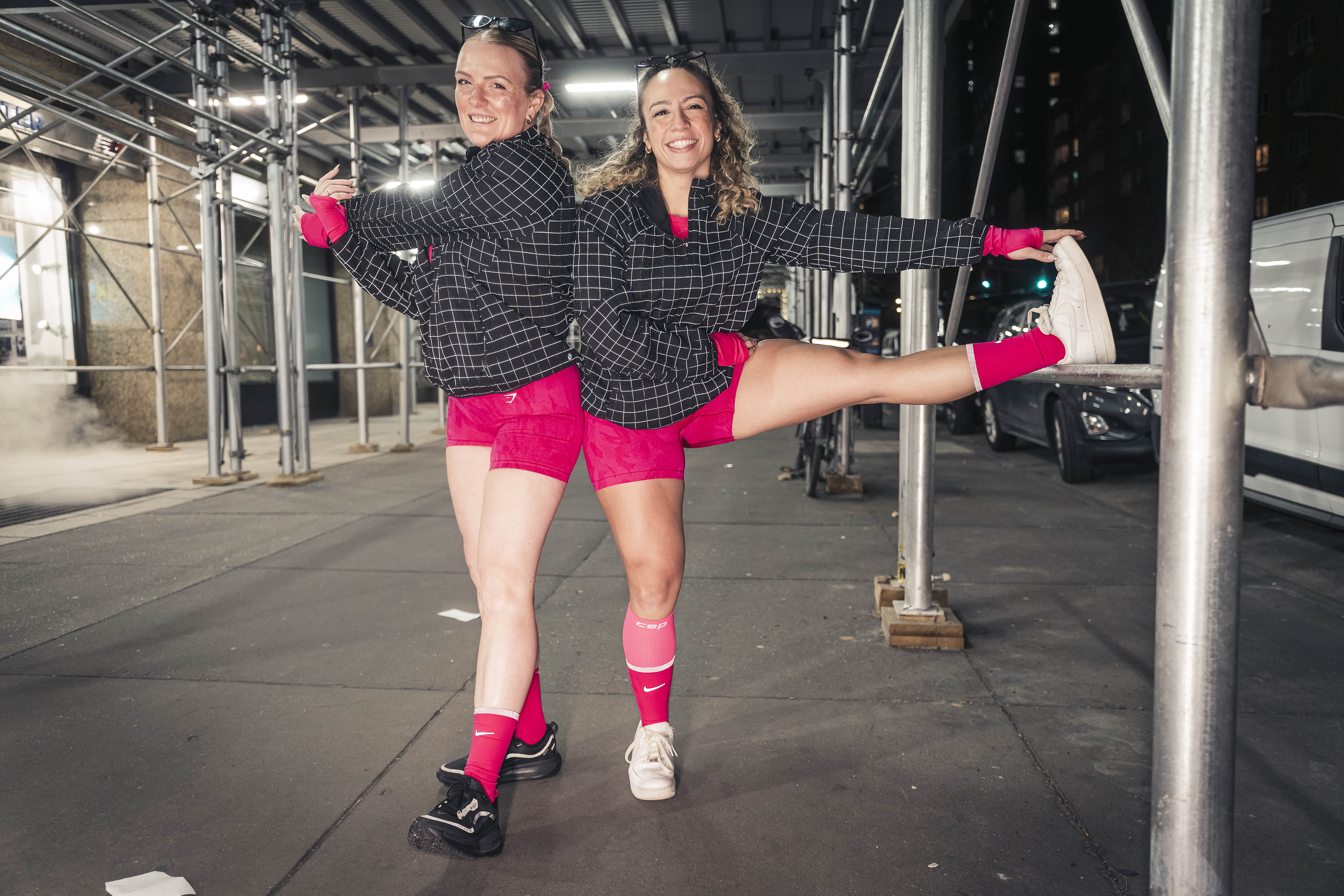 Runners Emily Heald and Becca Lopez, wearing matching pink and black outfits, stretching before the NY Road Runners Midnight Run.