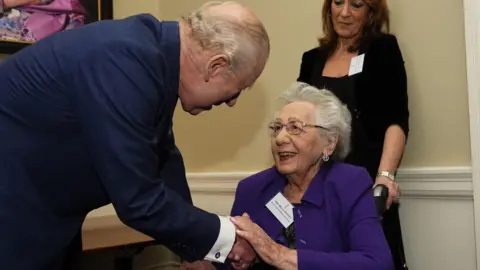 PA Media King Charles greets Helen Aronson. He wears a navy jacket and bends to shake her hand. Aronson wears a purple jacket, has short grey hair, and smiles at the King.