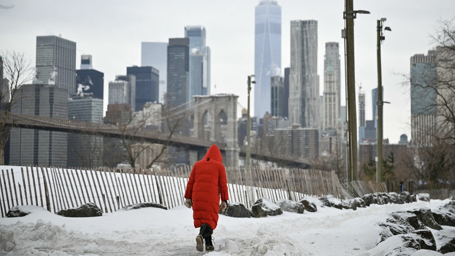 The Lower Manhattan skyline and Brooklyn Bridge are seen in the background as a woman walks through the snow in the Brooklyn borough of New York City on January 26, 2026. A monster storm barreling across the United States had killed at least 11 people on Monday, prompting warnings to stay off the roads, mass flight cancelations and power outages after a weekend of misery. The storm dumped snow, sleet and freezing rain across swathes of the country from Texas to New England, with temperatures set to fall dangerously low this week. (Photo by ANGELA WEISS / AFP via Getty Images)