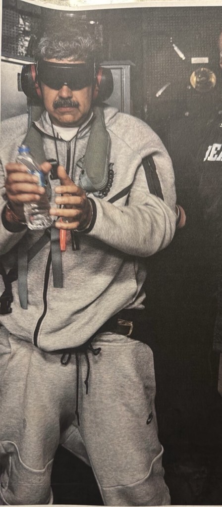 A man with a mustache wearing a blindfold, headphones, a grey hoodie, and grey sweatpants, holding a water bottle.