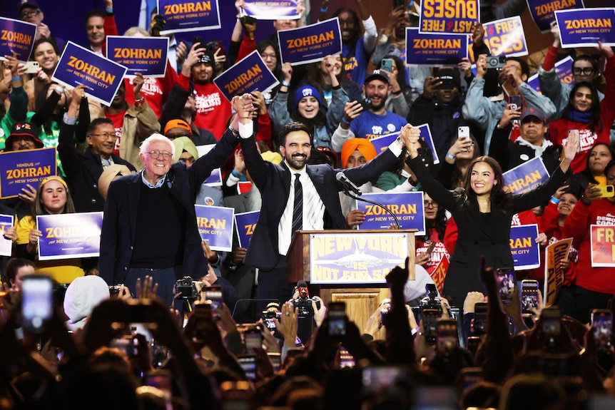 Zohran Mamdani raises his hands at a rally with Bernie Sanders and Alexandria Ocasio-Cortez