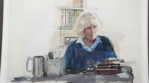 PA Media A portrait of Queen Camilla. She is sitting at a desk with a pile of books in front of her. There is a jar of pens and a tankard. She is wearing a blue stripy shirt and blue sweater. There is a book shelf in the background. 