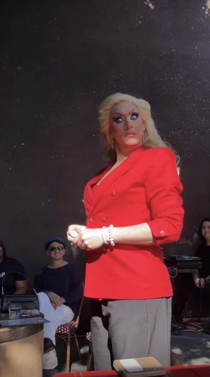 A drag queen dressed as Erika Kirk staring down the camera. She is wearing a bright red blazer and ice blue contacts.