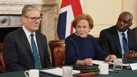 PA Media Sir Keir Starmer, Mala Tribich, and David Lammy, sit alongside each other at a green table in front of a union jack flag and a marble fireplace. 